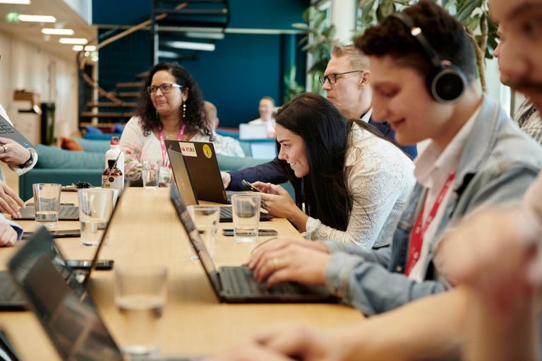 En gruppe mennesker sitter rundt et bord og jobber på bærbare datamaskiner. De ser konsentrerte ut, med noen som lytter med hodetelefoner, mens andre er dypt engasjert i diskusjoner. Miljøet ser ut som et moderne kontor med naturlig lys og en avslappet atmosfære.