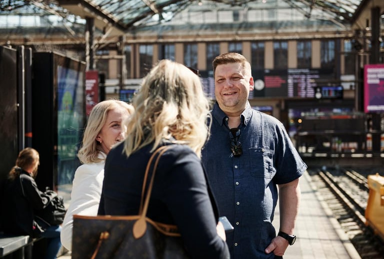Tre personer står sammen på en togstasjon, engasjert i en samtale. En smilende mann i blå skjorte og to kvinner er synlige, med togstasjonens travle bakgrunn.