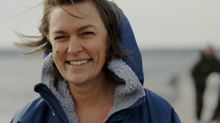 Smilende kvinne på vindfull strand ser inn i kamera